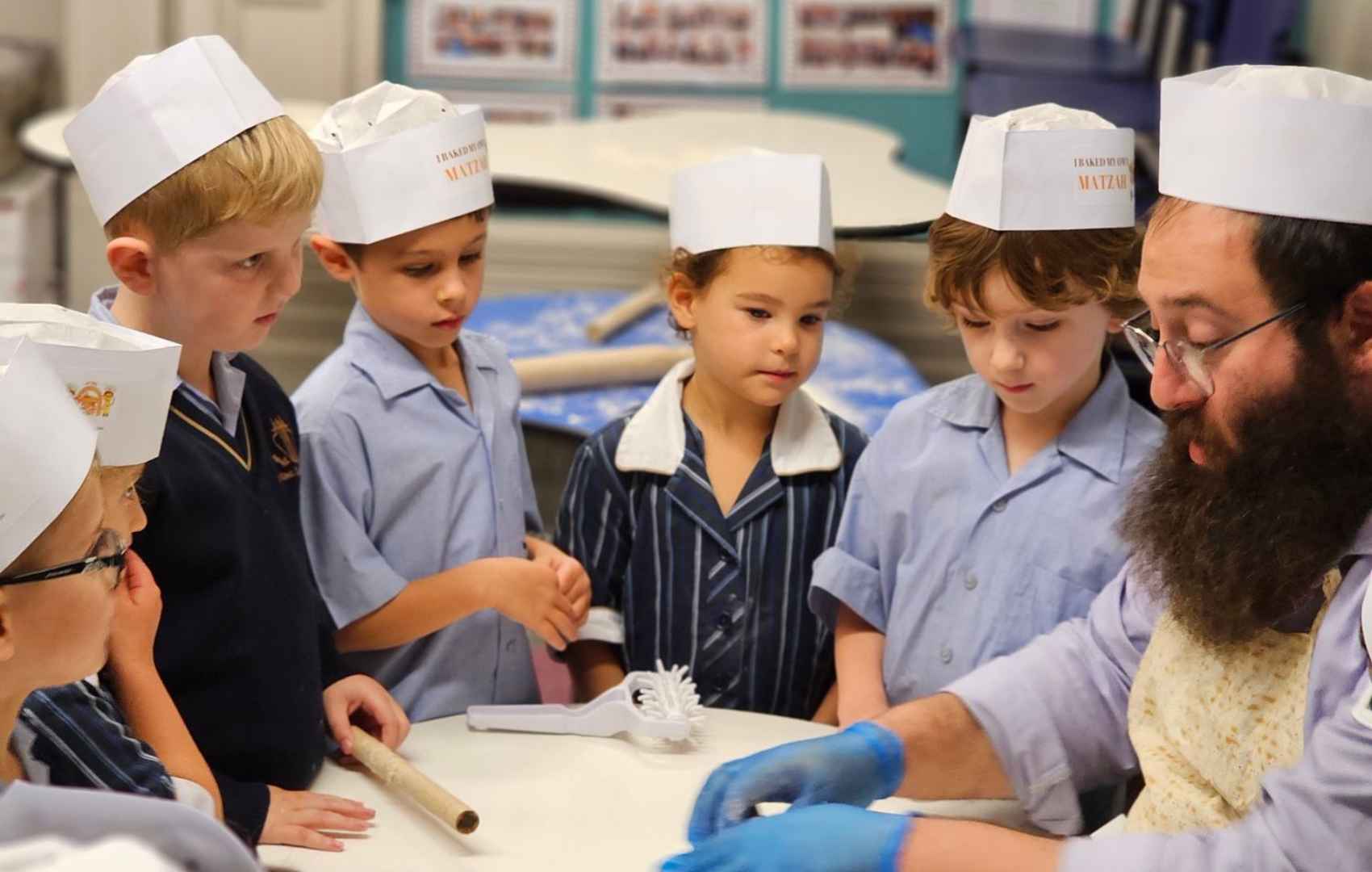 Six Prep students join Rabbi Yossi making matzah for Pesach. They are wearing white chefs hats with their school uniform.