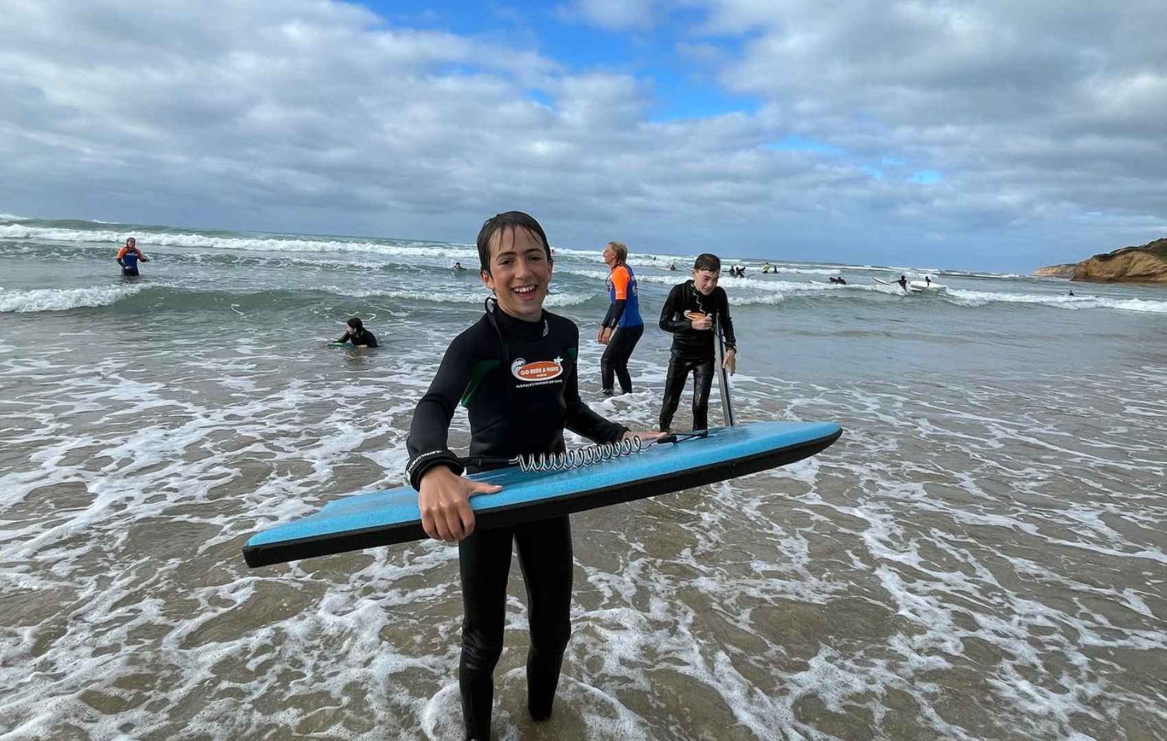 A Year 7 student is wearing a black wet suit and holding a blue body board. The student is standing in the ankle-deep sea, they have just been riding the waves.
