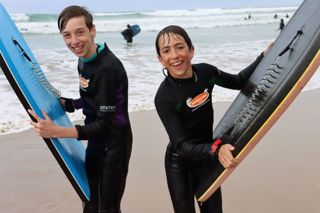 Two students are in front of the sea, they are holding bodyboards and wearing wetsuits. They have just come from the water. They are smiling and posing with their bodyboards.