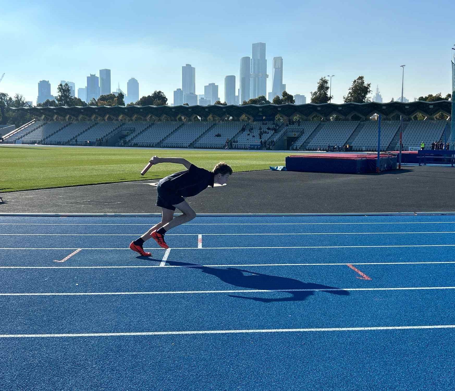 A senior school student is racing on a blue track. He is holding a baton and is participating in a relay. The Melbourne city skyline can be seen behind him.