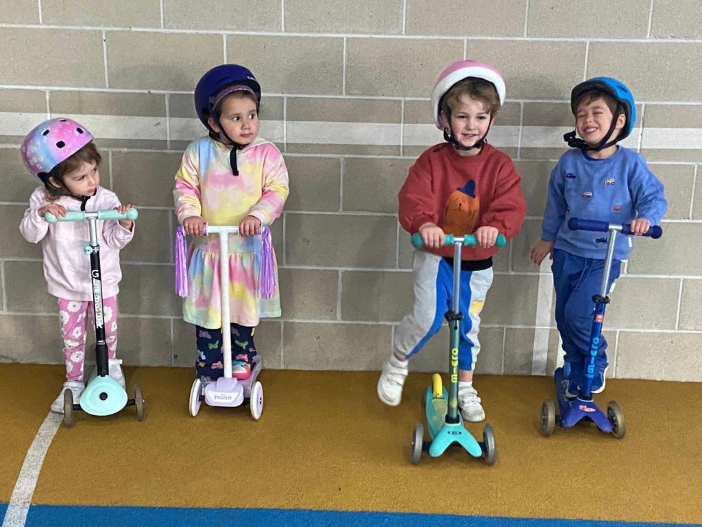 Four ELC students pose with their scooters. They are wearing helmuts and are excited about being able to scoot