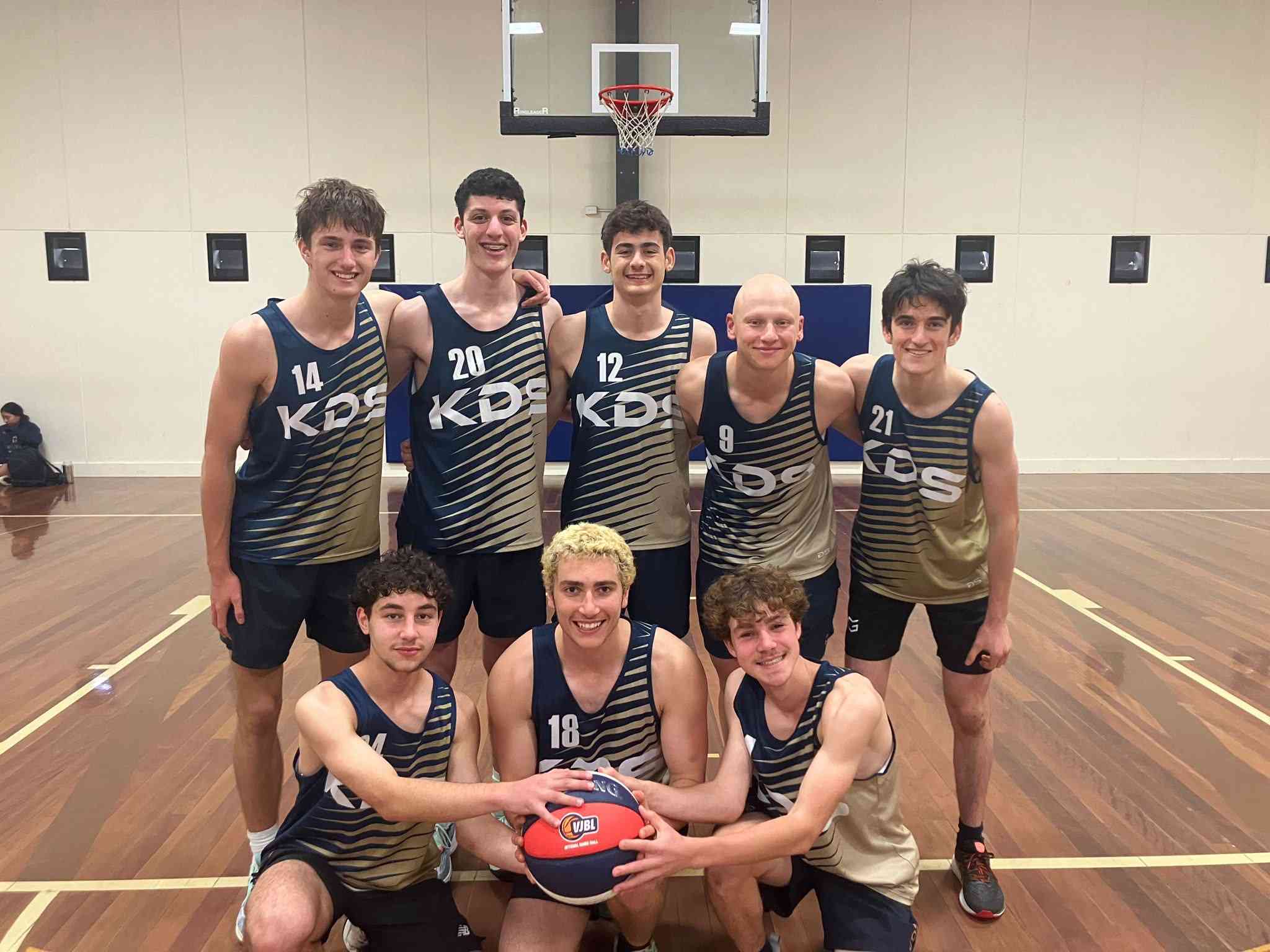 Senior boys basketball team 2024 huddle together with a ball in the middle. They are looking at hte camera and smiling. They have made it to the season final.