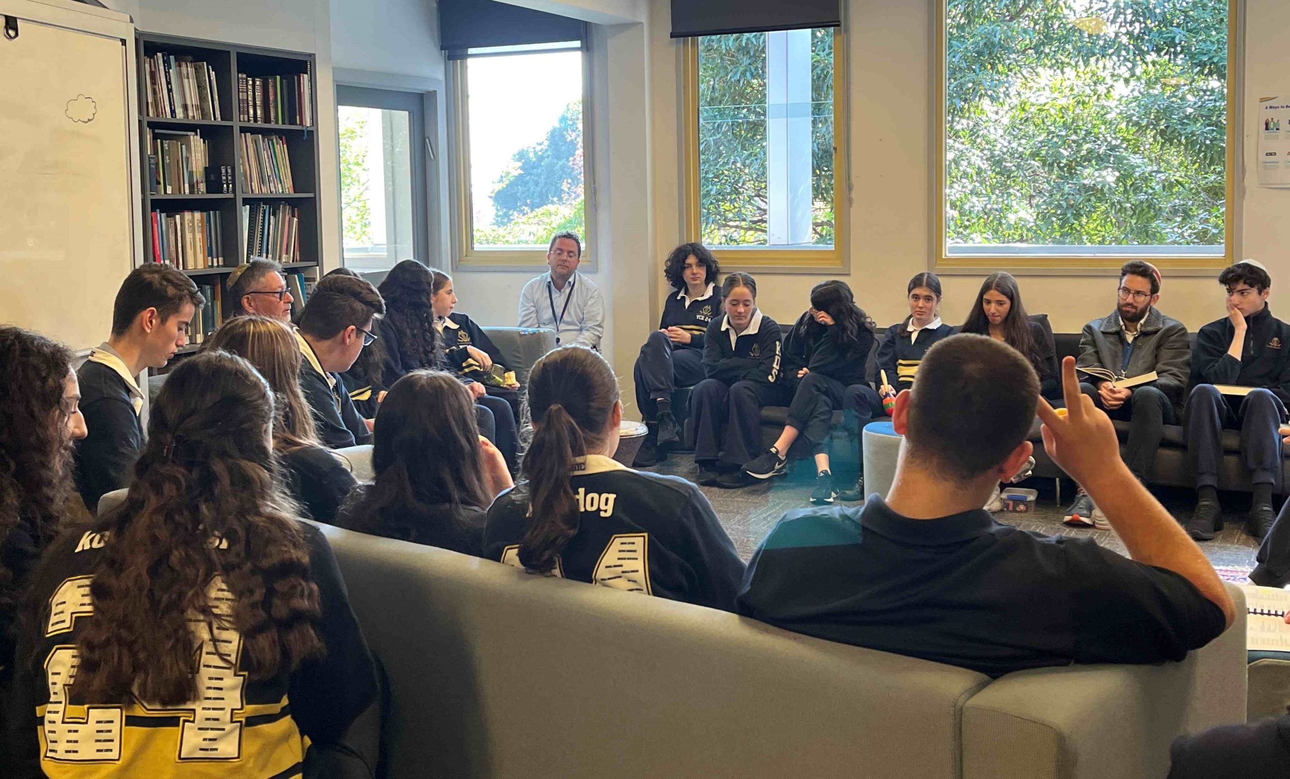 Students are in the J-Life room, singing and listening as part of preparing for Shabbat with T'filah - prayer. They are wearing school uniforms.