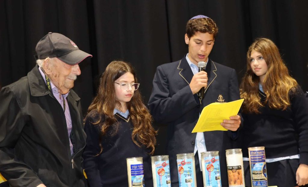 Three King David students are one Holocaust survivor stand in front of six memorial candles commemorating Yom HaShoah. One student is holding a yellow peice of paper and talking into a microphone. They are sombre