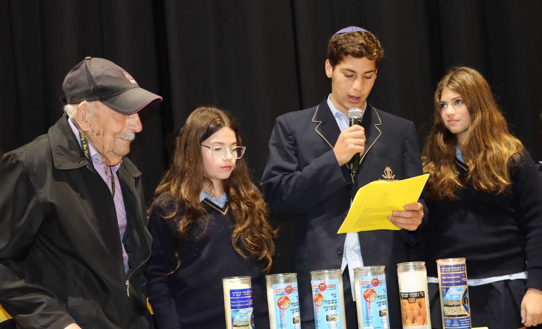 Three King David students are one Holocaust survivor stand in front of six memorial candles commemorating Yom HaShoah. One student is holding a yellow peice of paper and talking into a microphone. They are sombre