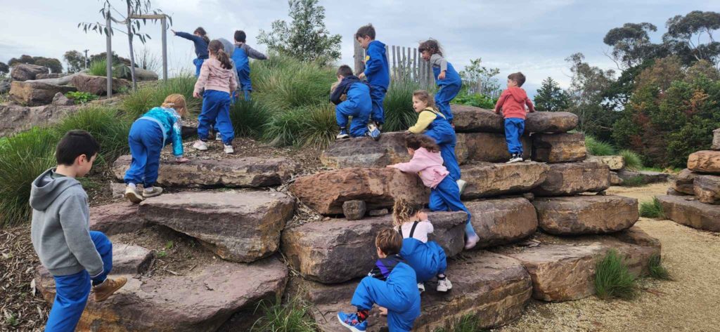 ELC Students are at Bush Kinder - they are wearing blue overalls for play and climbing on rocks.