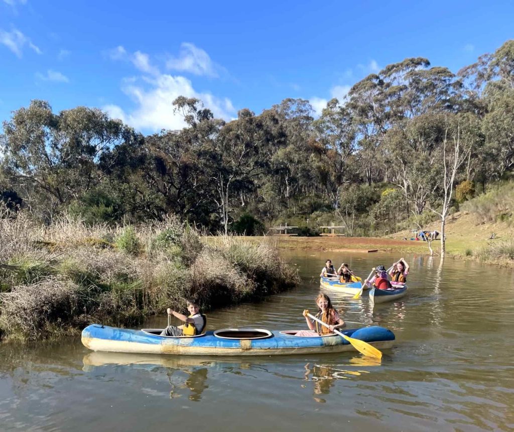 Year 5 students are in blue kayaks on water, using yellow oars. They are smiling and enjoying the sunny day on their adventure.