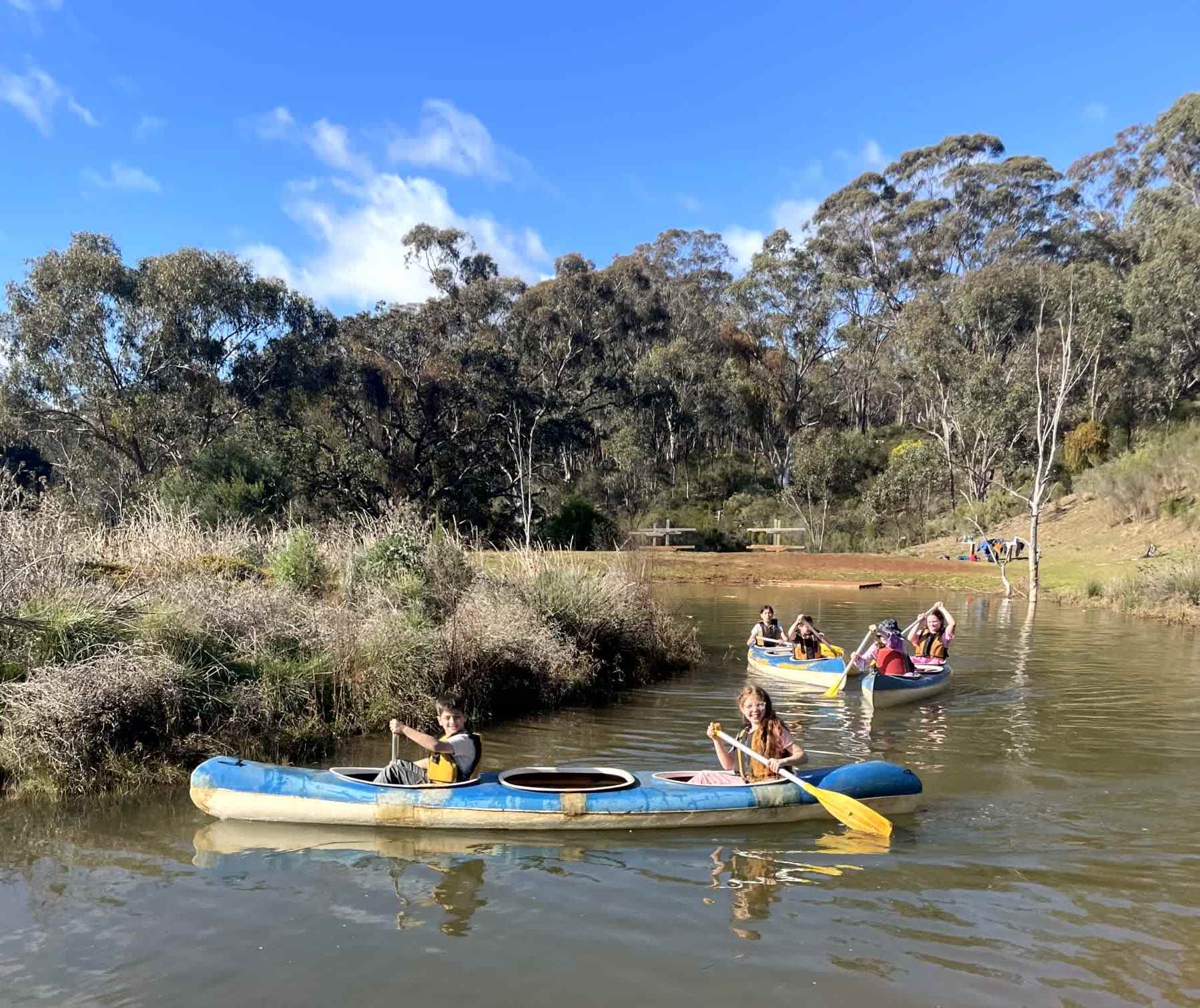 Year 5 students are in blue kayaks on water, using yellow oars. They are smiling and enjoying the sunny day on their adventure.