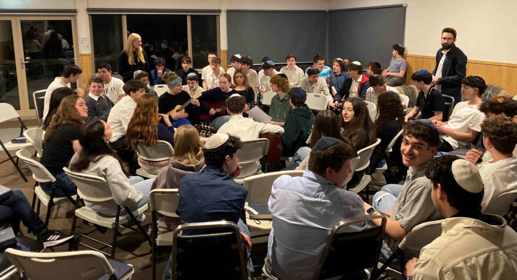 Students are celebrating Shabbat at our Year 10 Shabbaton. They are sitting in a circle, singing as a student plays guitar.