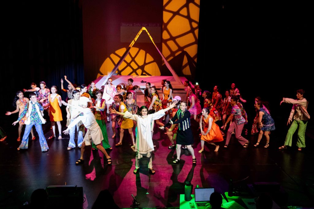 Years 6-9 students are on stage, dancing in front of a pyramid. They are wearing colourful costimes and are performing in the musical Joseph and the Technicolour Dreamcoat.