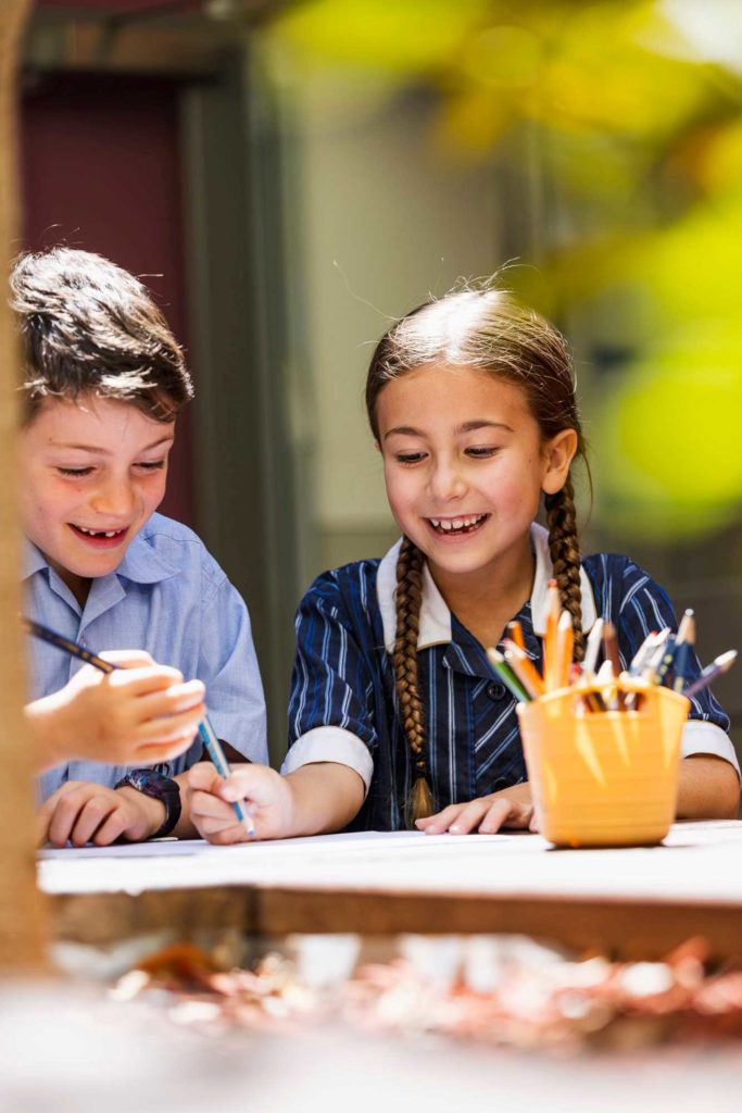 Two Junior School students are excitedly drawing. They are looking down at their work. There are coloured pencils in front of them and they are wearing school uniform.