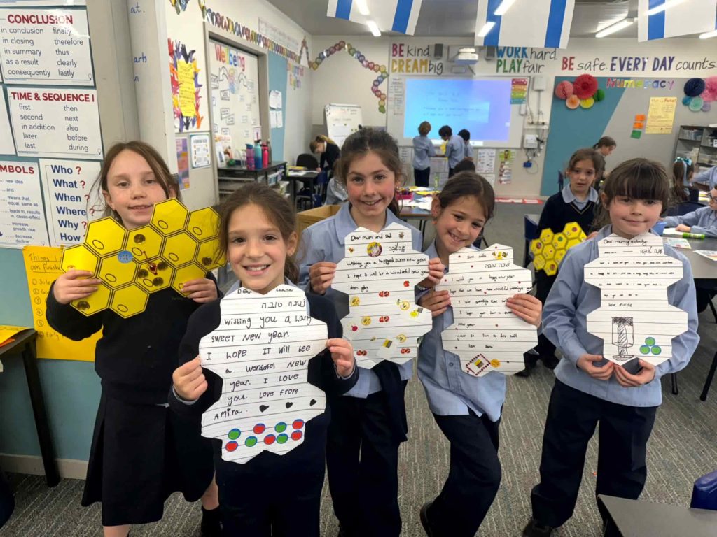 Five students are in school uniform. They are smiling and holding up their Rosh HaShanah work that is in the shape of a beehive.