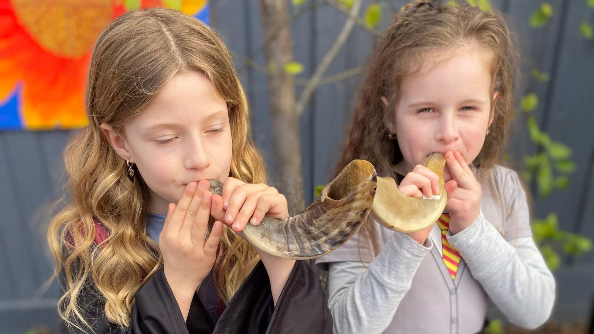 Two Year 2 students sound the Shofar. They are holding a shofar in their mouths and blowing. They are preparing for Elul.