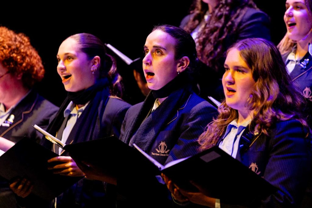 Six young people are wearing King David academic uniform. They have their mouths open and are looking outwards, not at the camera. They are singing in the 2024 Spring concert.