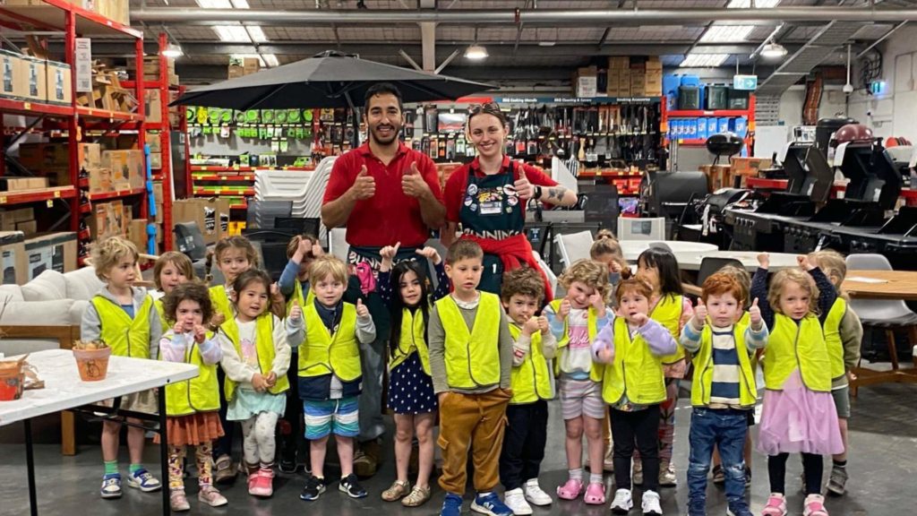 Gan Gefen are at Bunnings. They are wearing yellow safety vests. They are together, looking at the camera and smiling, They have just made 'bug hotels' - safe small houses for bugs and plants to thrive.