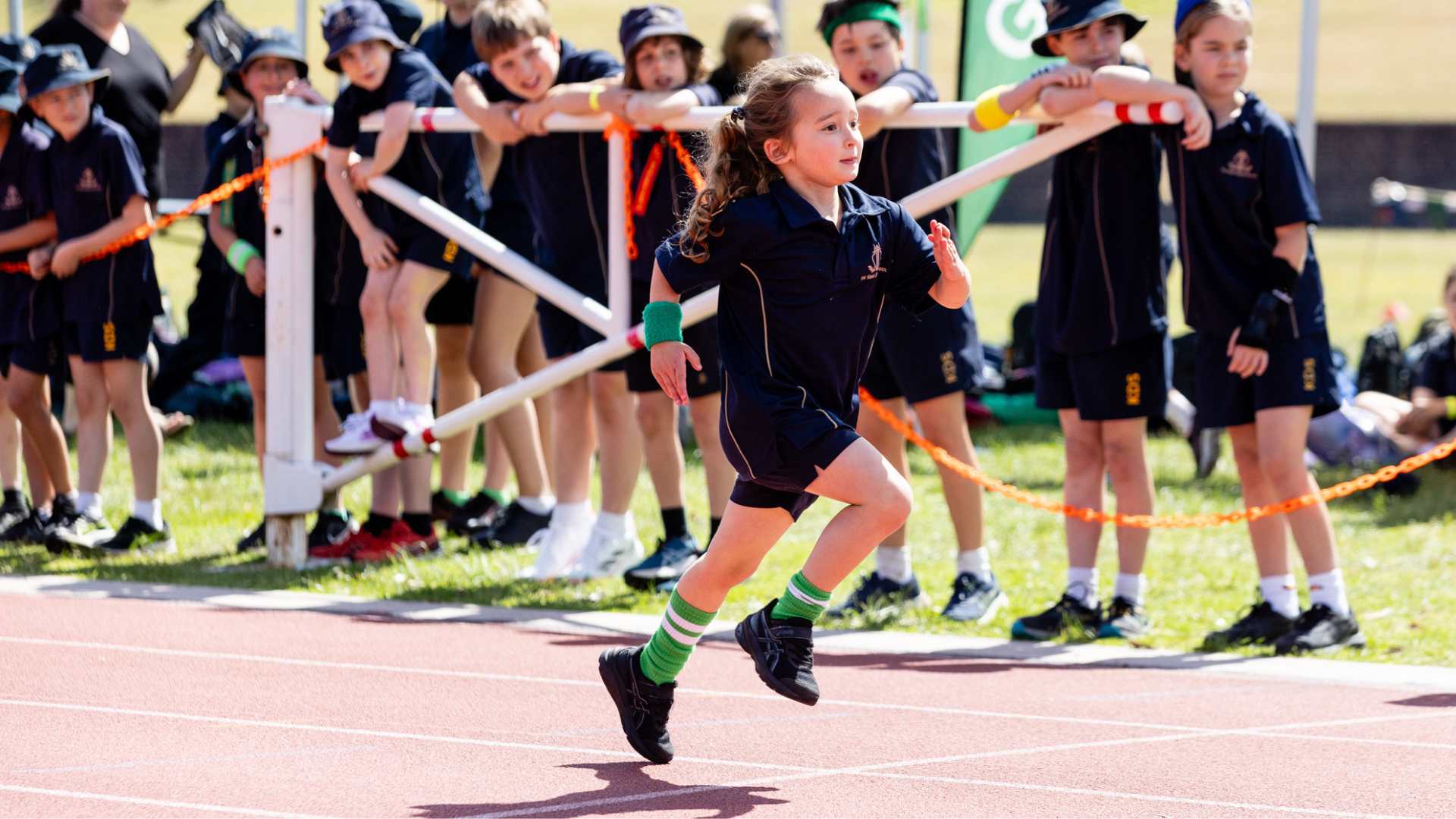 A Junior School student is running on an athletics track, being cheered on by other students. She is wearing The King David School uniform and has green accents for her sports house, Galil