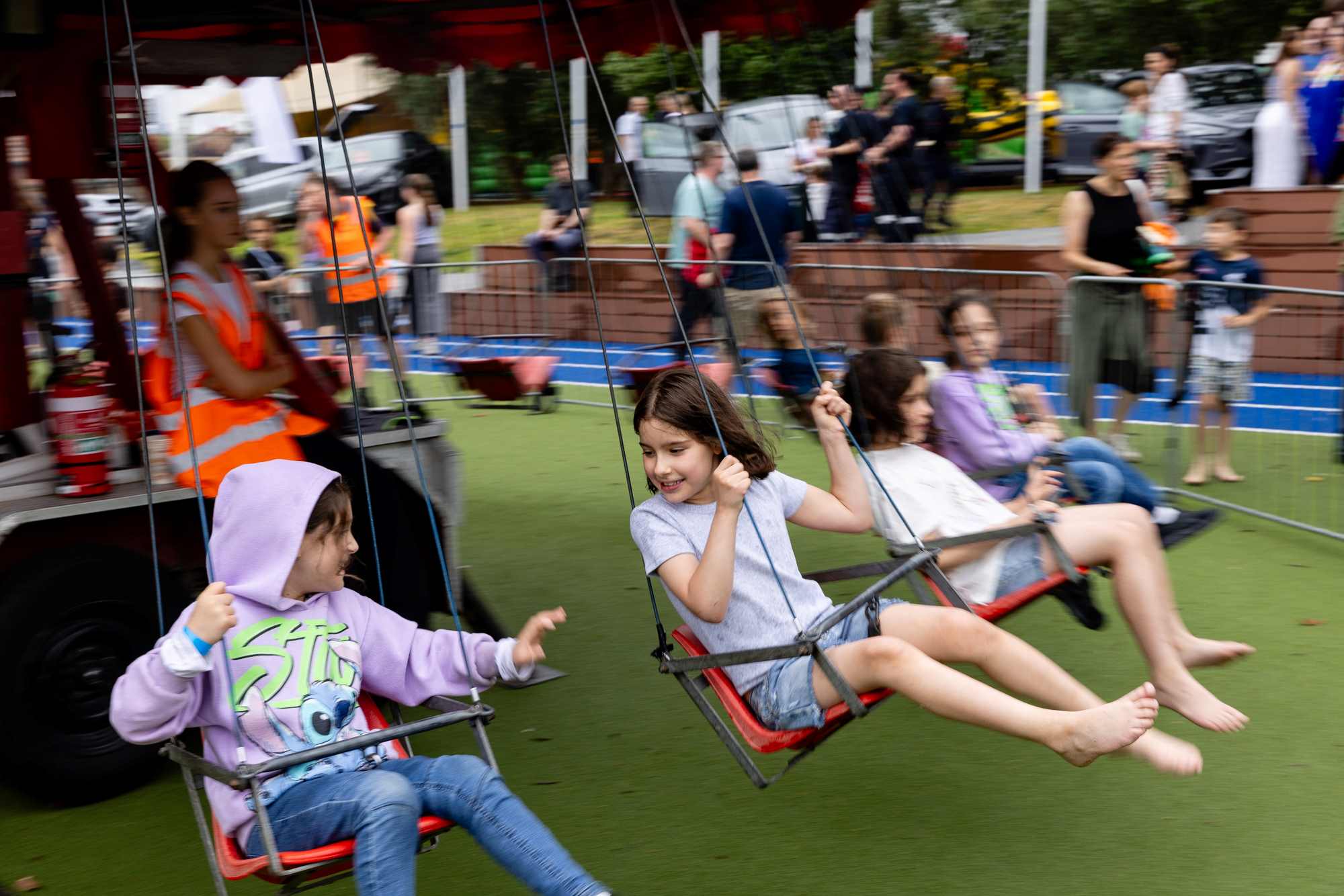 Four girls dressed in casual clothes - shorts and hoodies - are riding the chair-o-plane at the King's Carnival. They are swinging in chairs off the ground. They are happy.