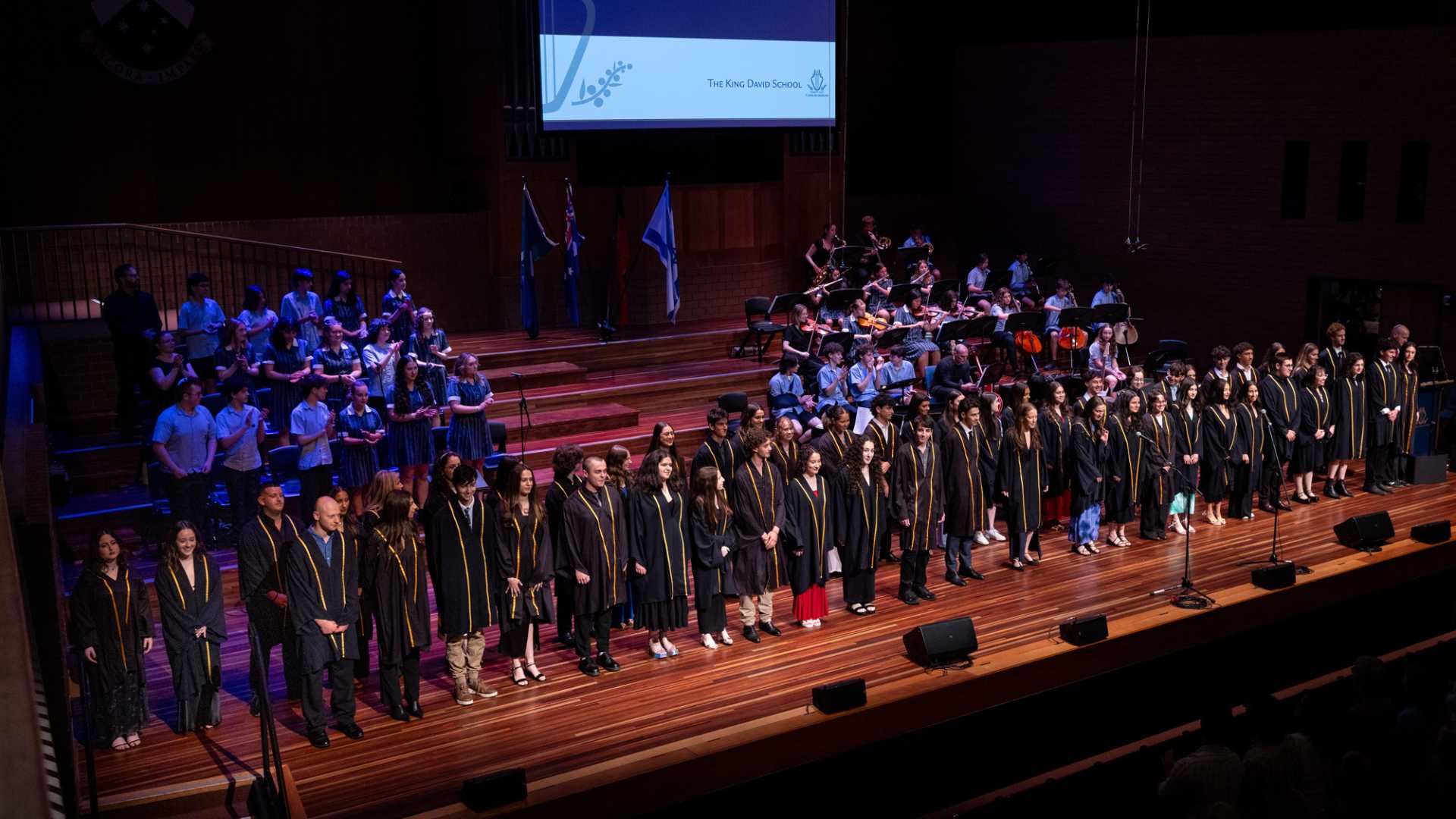 The Class of 2024 is on stage at Presentation Evening. They are wearing their graduation robes. Student musicians are also on stage, singing and palying musical instruments.
