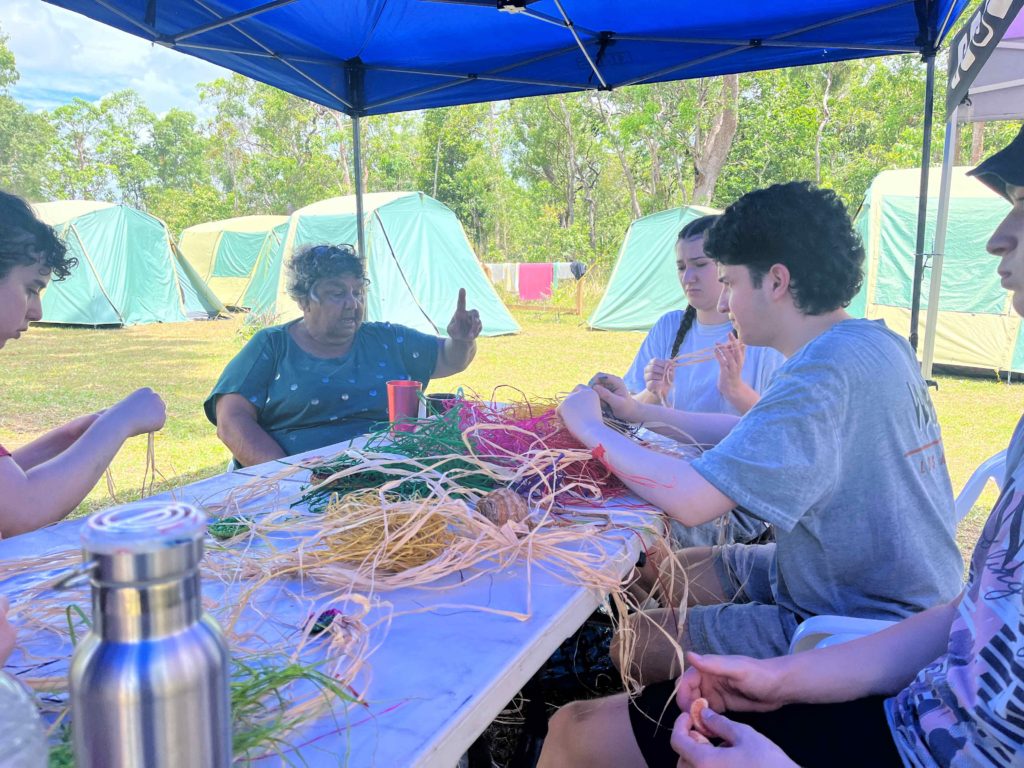 Traditional Owner Aunty Esme tells Year 11 students on camp about working as a domestic servant. They are on the Gumbiir homeland. They are using colourful rattan to learn how to weave.