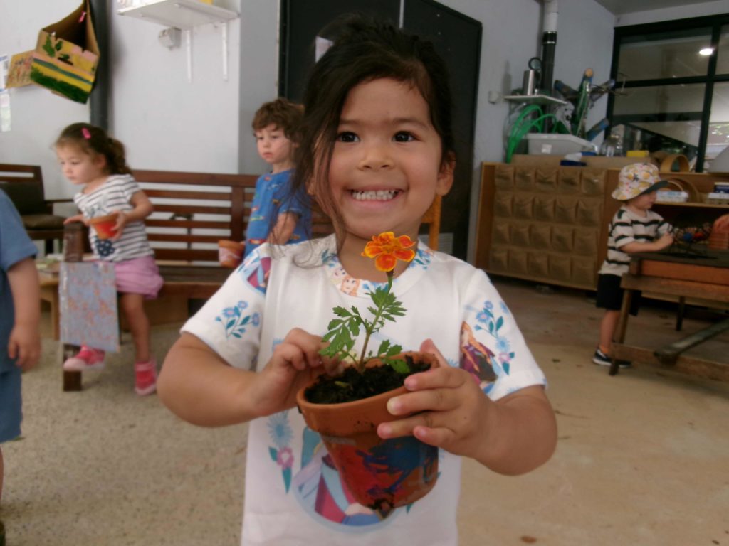 A Junior Kinder student is holding a pot with an orange flower she has just planted. She is happy. She is celebrating Tu BiSh'vat