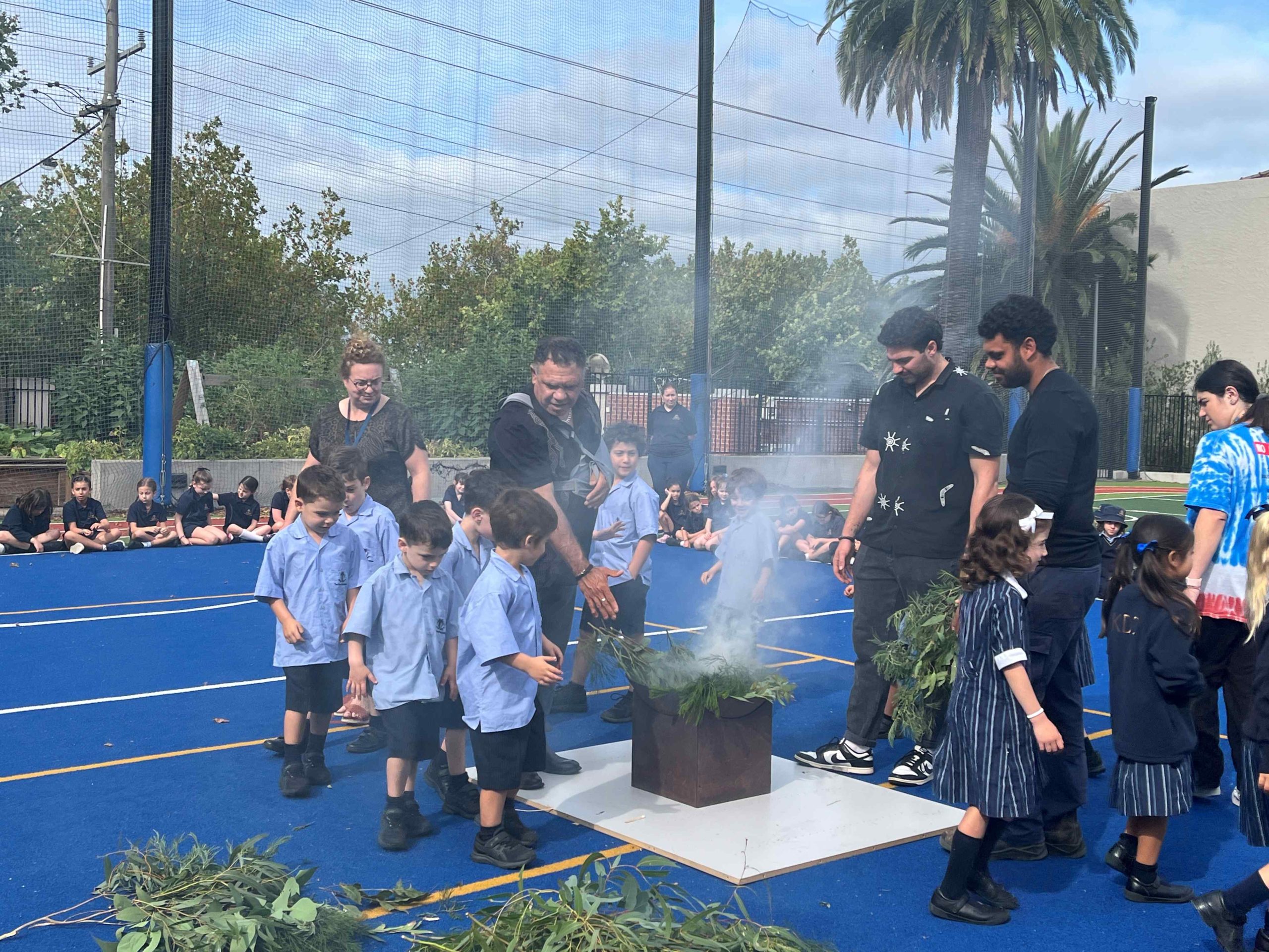 Junior School students are in school uniform, on the oval at a Welcome to Country smoking ceremony. They are walking through smoke in a ceremony led by William Pepper for the Boonwurrong Land and Sea Council.