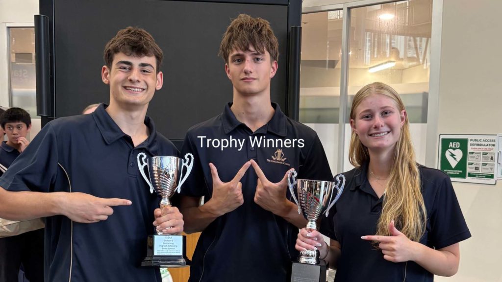 Three Senior School students are smiling, two are holding chrome trophies, one is signalling 'winner' with his hands. They are wearing navy sports uniforms. they are happy because we have come 3rd place in the EISM Swimming Carnival.