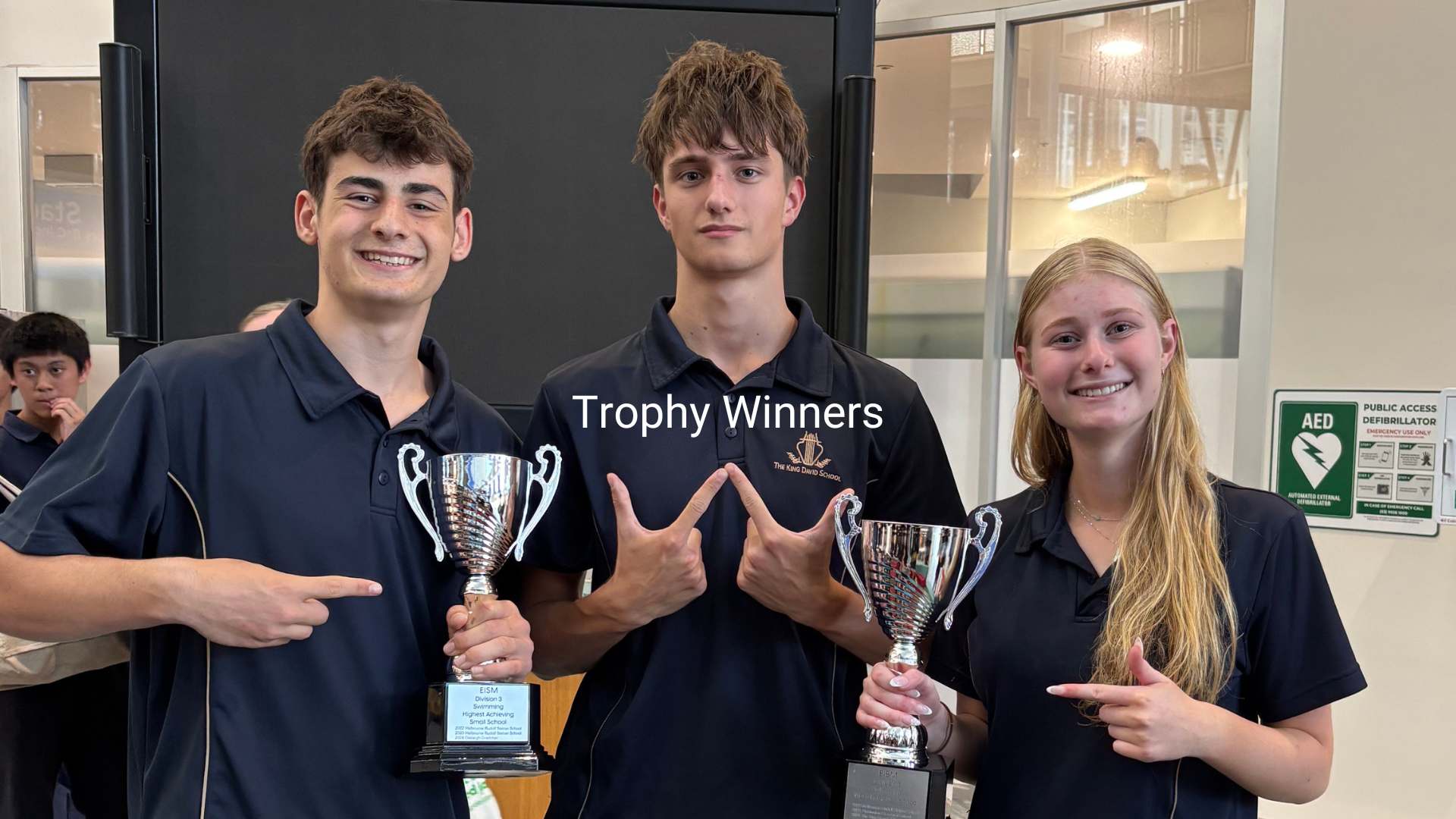 Three Senior School students are smiling, two are holding chrome trophies, one is signalling 'winner' with his hands. They are wearing navy sports uniforms. they are happy because we have come 3rd place in the EISM Swimming Carnival.