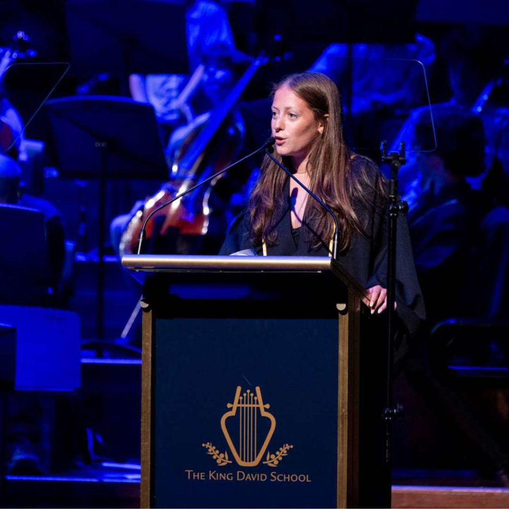 Class of 2024 graduate Poppy is speaking at Presentation Evening. She is speaking into a microphone at a King David School branded lectern. She is happy to be graduating.
