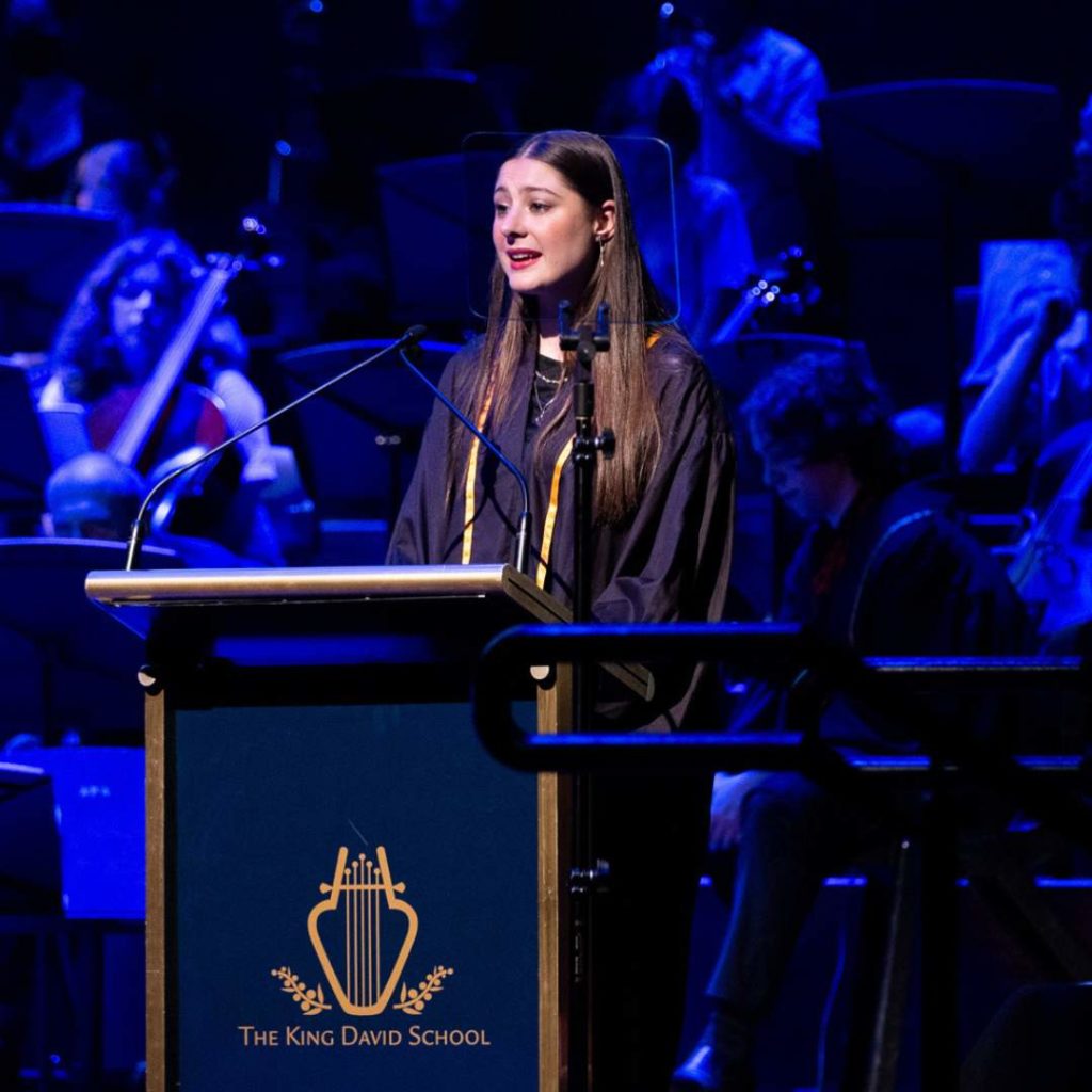 Class of 2024 graduate Trixie is speaking at Presentation Evening. She is speaking into a microphone at a King David School branded lectern. She is happy to be graduating.
