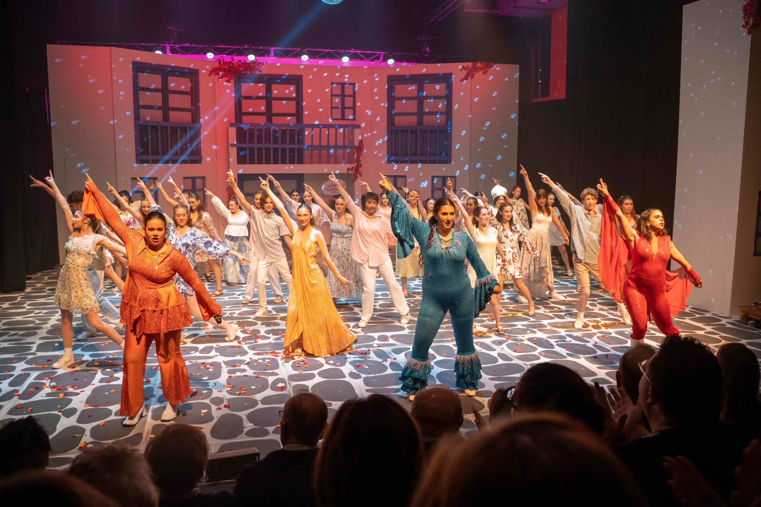 Students are on stage in 1970s stylee costumes. They are dancing and performing in the finale of Mamma Mia the musical.