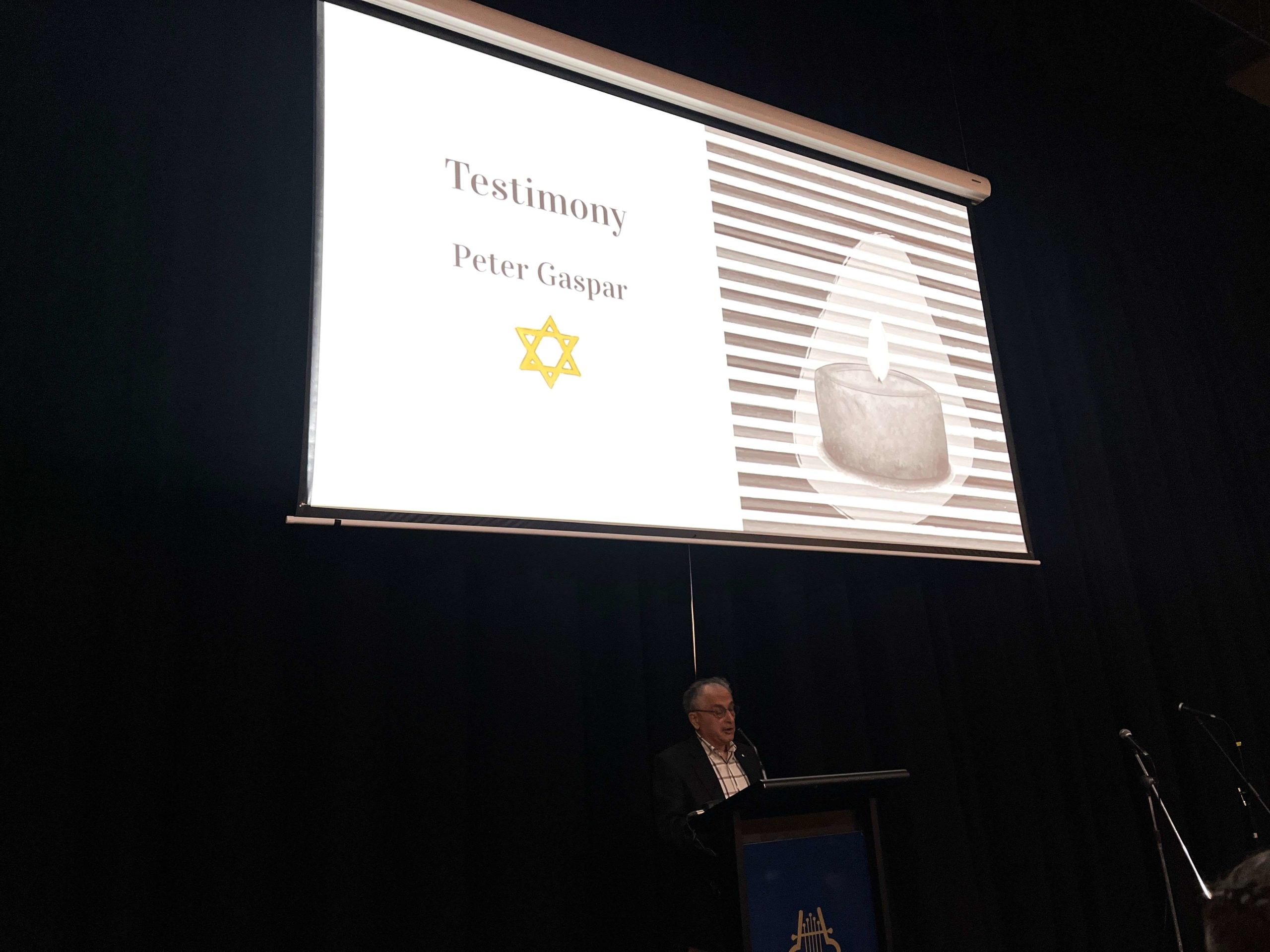 Shoah survivor Peter Gaspar speaks at King David's Yom HaShoah commemoration. He is standing in front of a slide that says 'survivor testimony'.