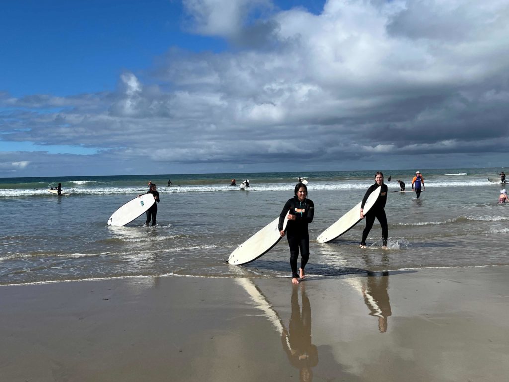 Year 7 students are emerging from the sea in wetsuits with surf boards.