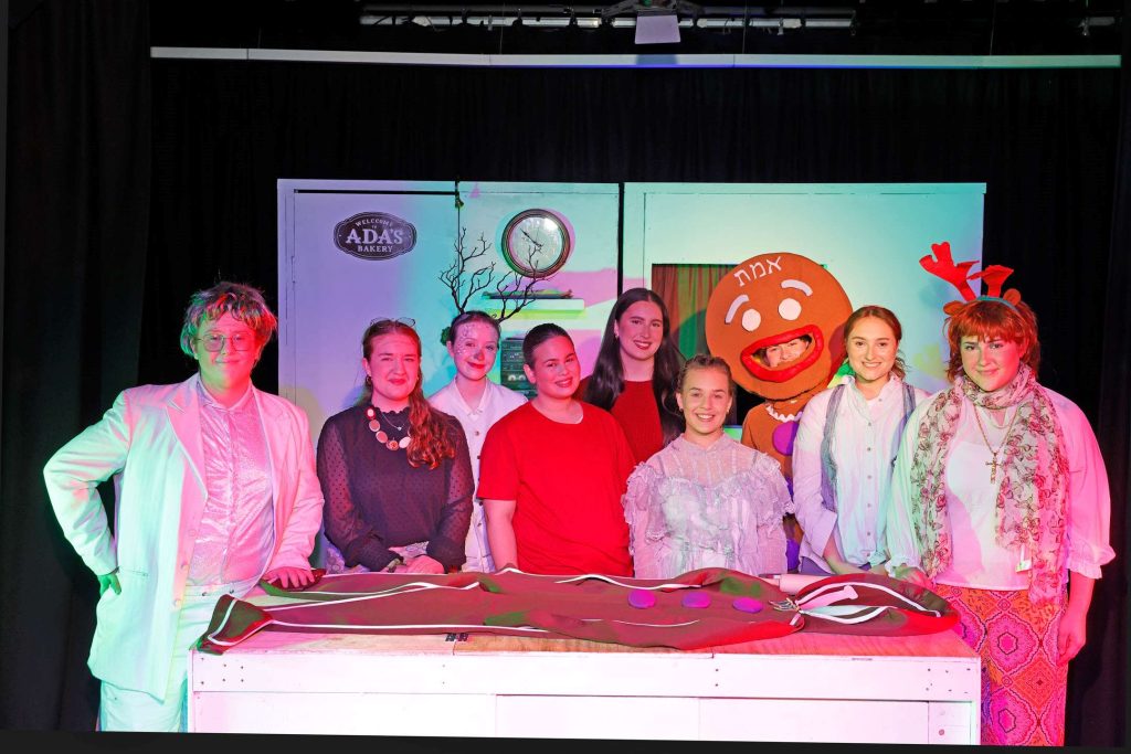 The cast of a Very Jewish Christmas Carol are in costume, standing in front of a Jewish bakery kitchen. They have just performed Elise Heart's play.