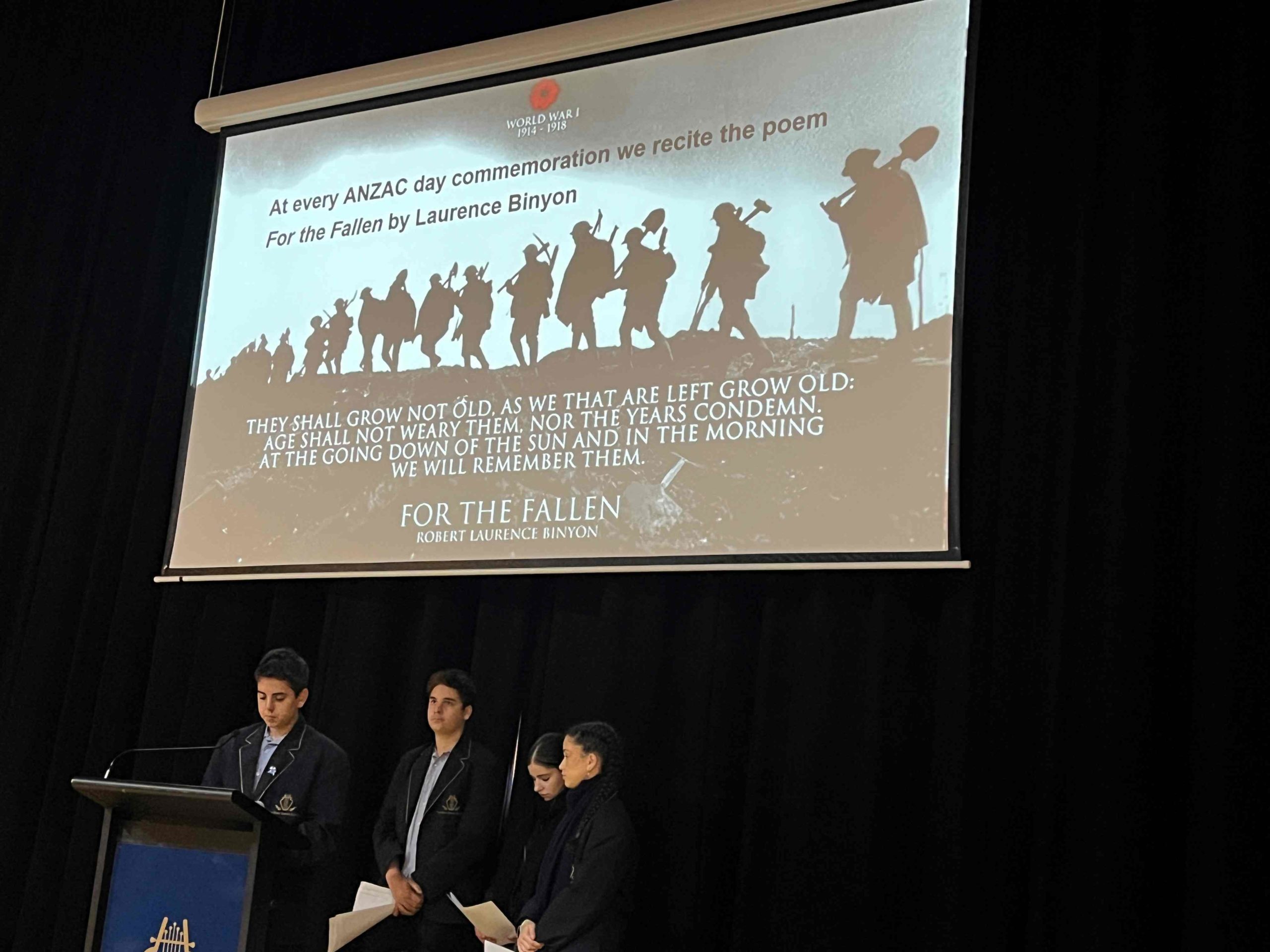 Students stand in front of a presentation of Laurence Binyon's poem 'For the Fallen'. They are wearing academic uniform. They are commemorating ANZAC Day at assembly.