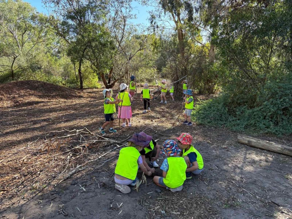 Kindergarten children in fluoro yellow vests play on the ground at Gan Tevah - Bush Kinder. They are surrounded by trees and excited to be in nature.