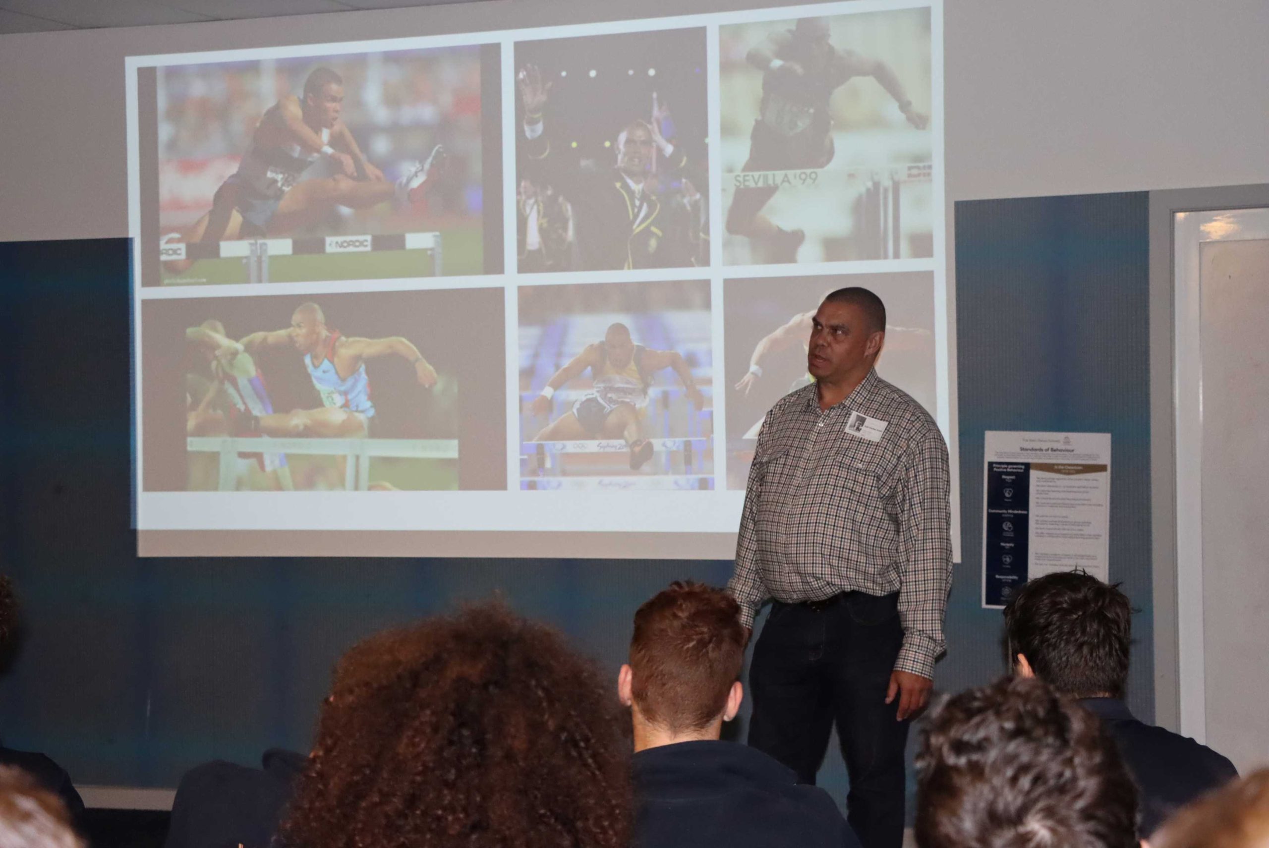 Kyle Vander-Kuyp speaks to students at the Magid Campus for Reconciliaton Week. He is standing in front of photos of his sports achievements.