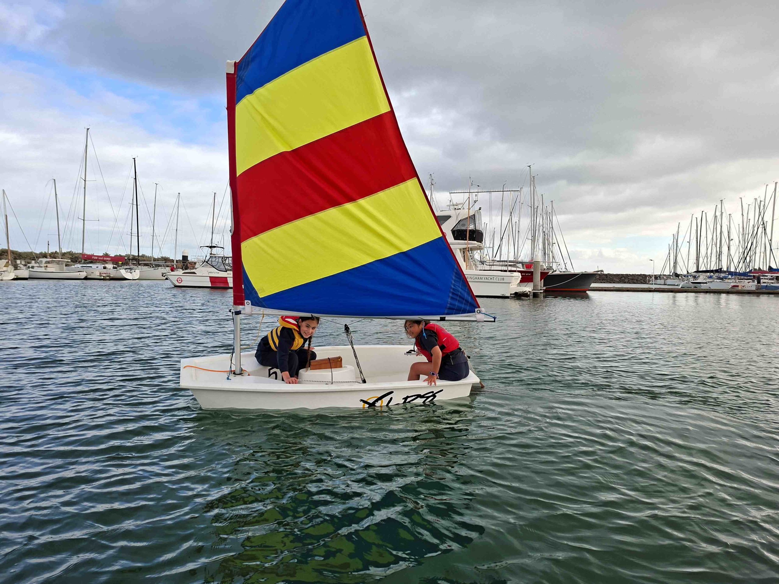 Year 4 students sail at Sandringham Yacht Club, with a yacht that has a yellow, blue and red striped sail. The water is smooth.