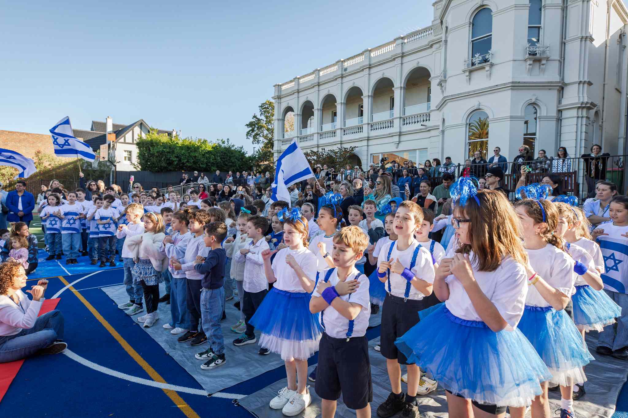 Students at the Junior School celebrate Yom HaAtzmaut on the oval. They are wearing blue and white - some are wearing blue ballerina tutus. You can see the school mansion behind the students.