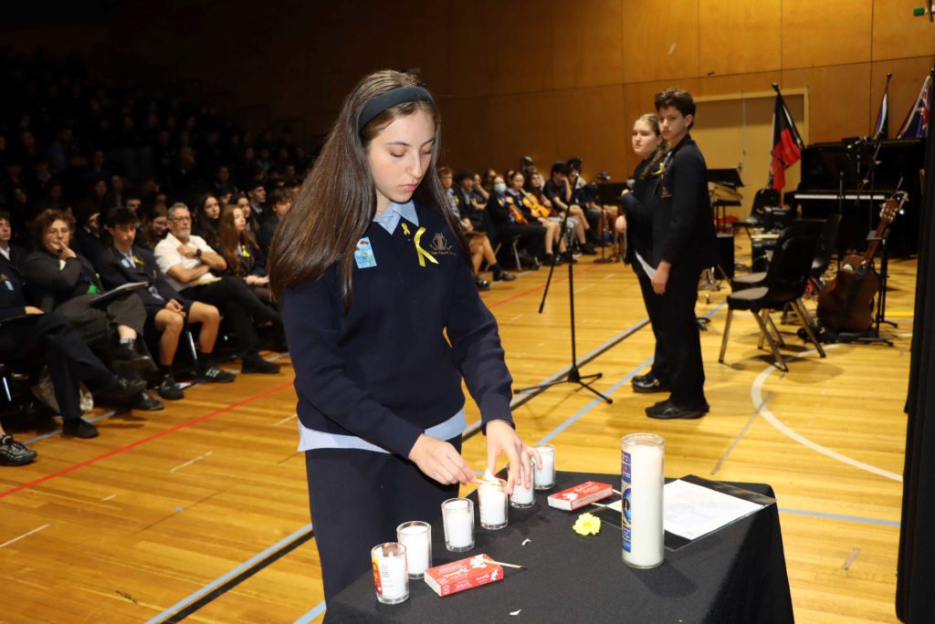 Students are commemorating Yom HaZikaron at the Magid Campus. One student is lighting a memorial candle while other students are standing at microphones during the assembly.