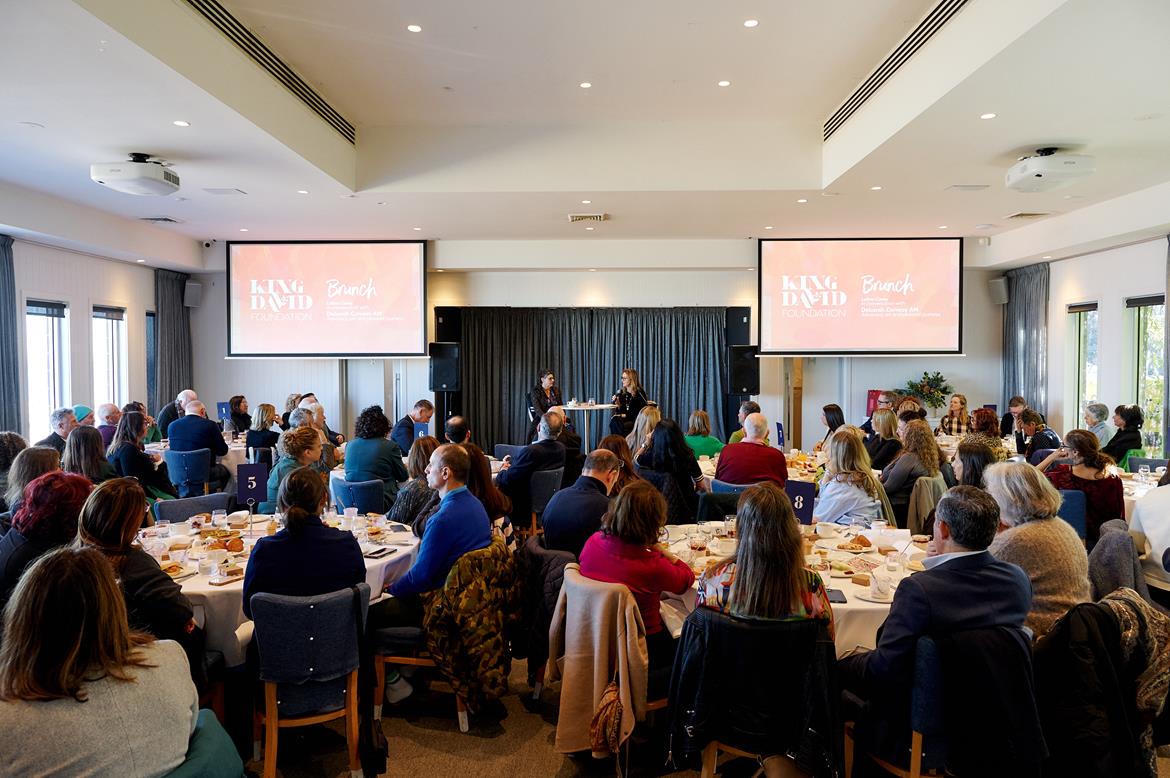 This shows a full room of people sitting at tables. There are two screens that will show photos. The people are attending a brunch to raise money for The King David School.