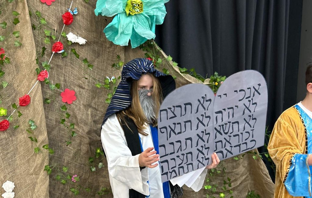 A student is dressed as Moshe receiving the 10 Commandments. She is holding the tablets with the Commandments on them. she is standing next to a fake Har Sinai, decorated with paper flowers.