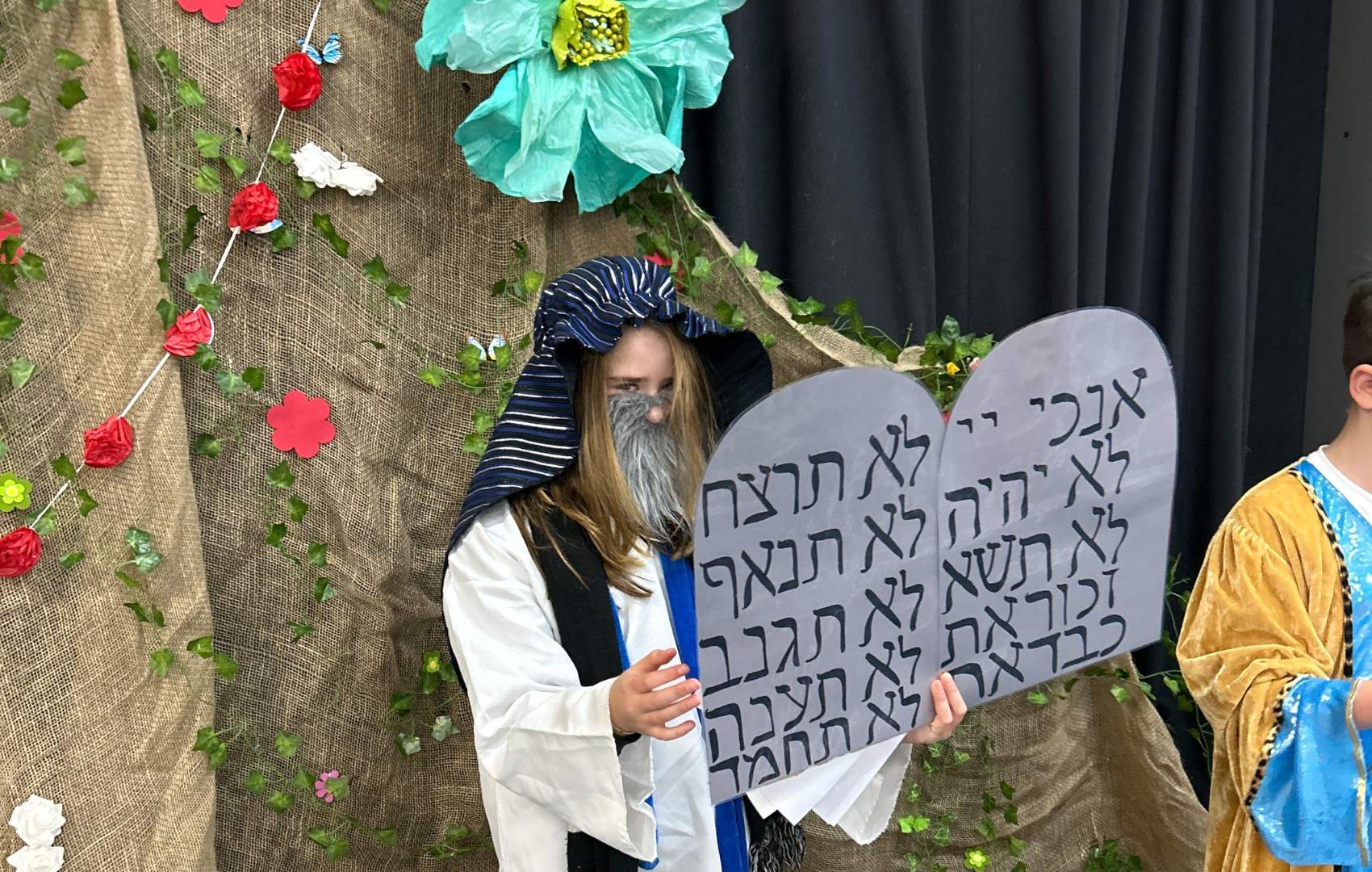 A student is dressed as Moshe receiving the 10 Commandments. She is holding the tablets with the Commandments on them. she is standing next to a fake Har Sinai, decorated with paper flowers.