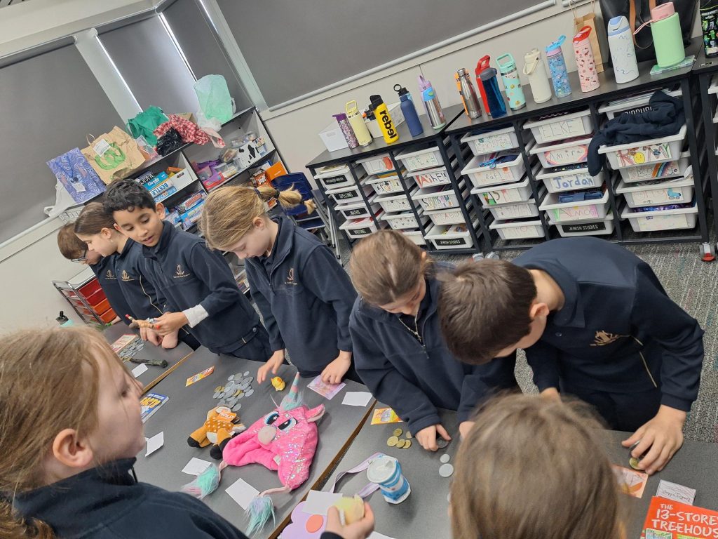 Year 3 students are buying and selling at the market that they made. They are looking at merchandise on a long table. They are wearing their school uniforms.