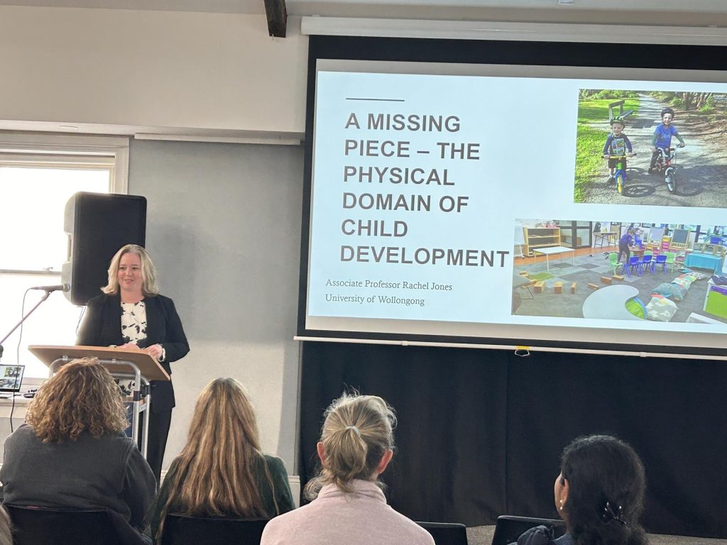 Presenter Dr Rachel Jones is speaking at a lectern. Behind her is a screen with a picture of children's playgrounds. She is presenting on the importance of movement in children's learning.