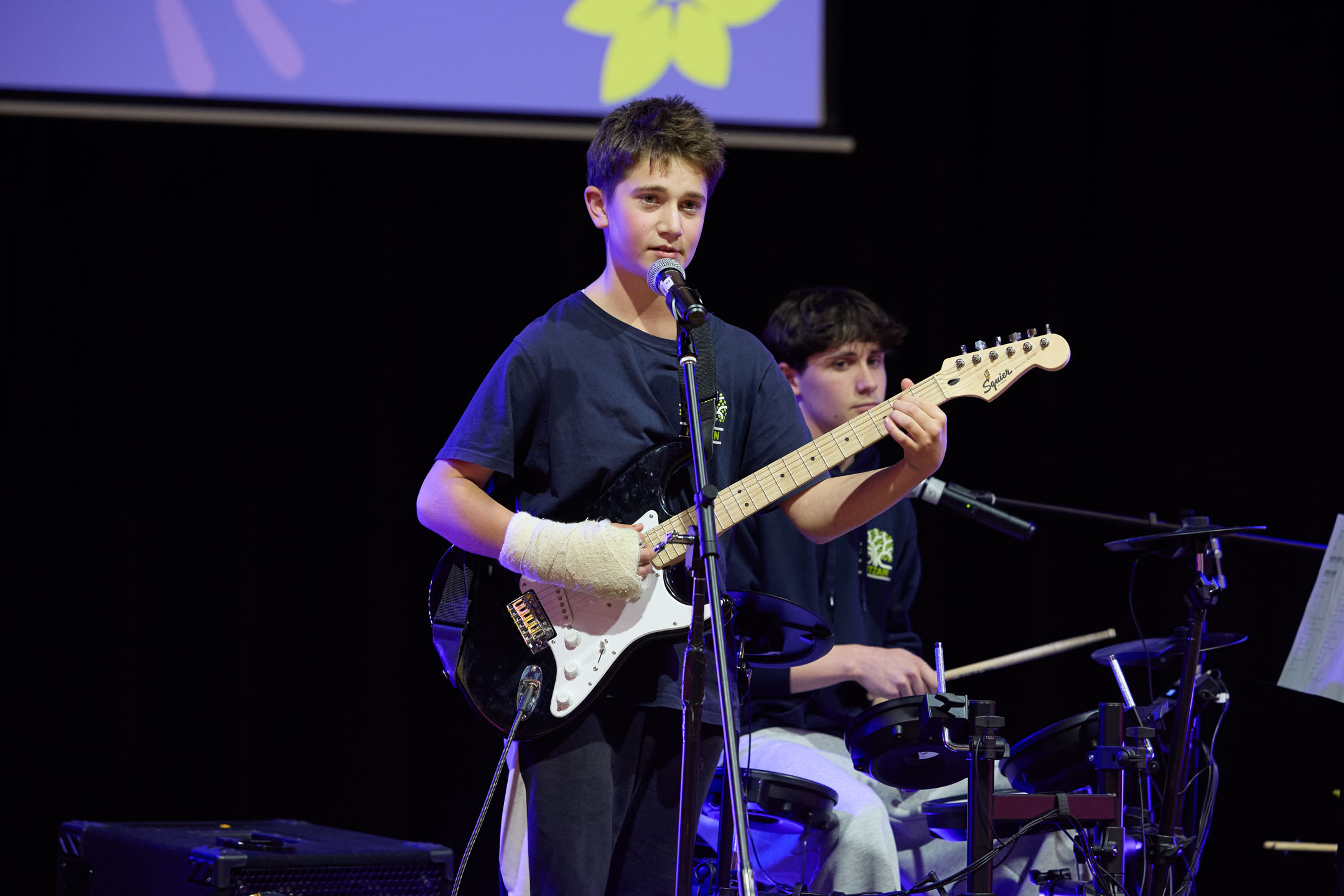 Year 9 student plays the guitar and sings while another student behind me plays the drums. They are on stage performing as part of Artivism - a showcase of Year 9 learning about art and change.