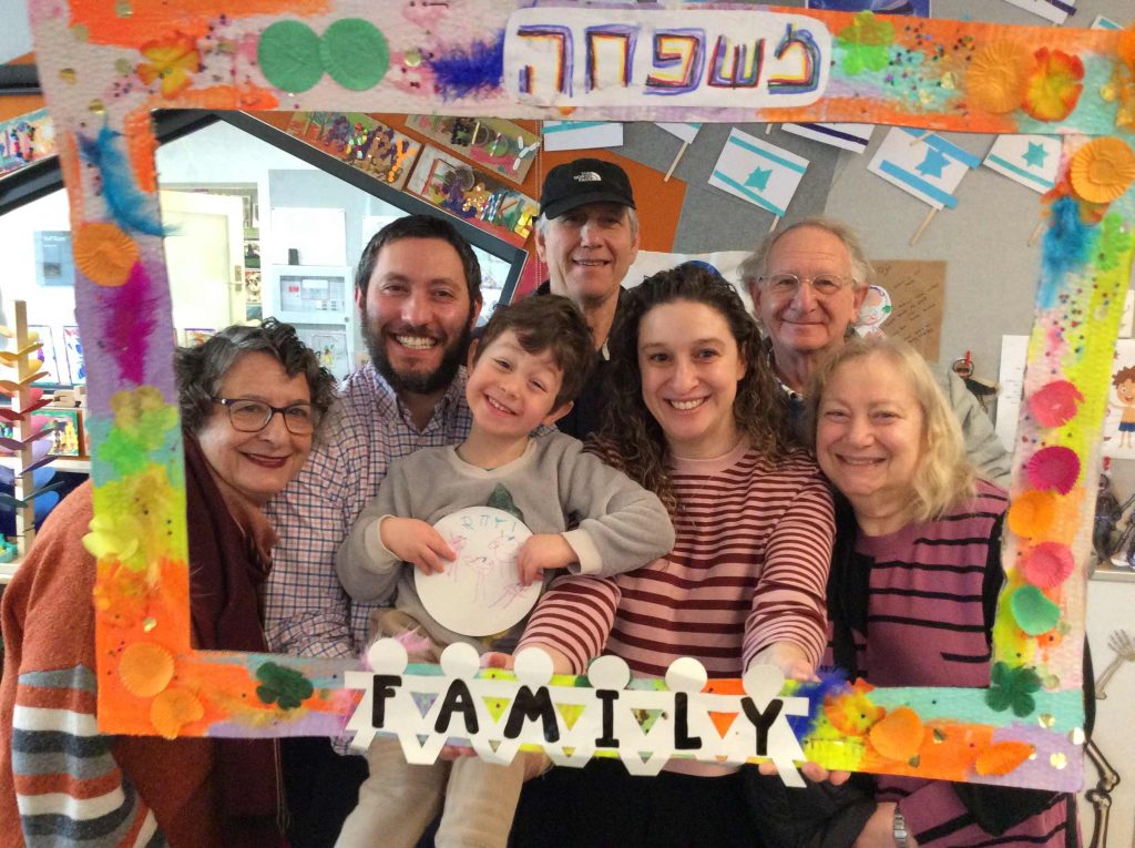 Seven family members of three generations are celebrating Family Day at our ELC. They are under a hand painted cardboard frame that says Family.