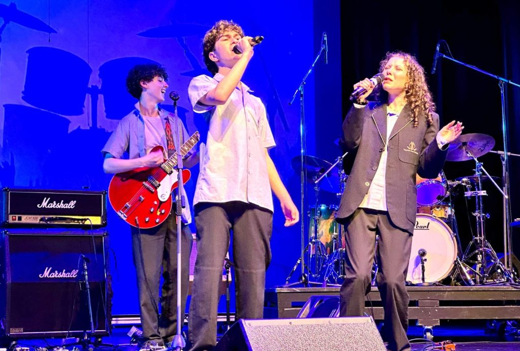KDS students are performing in a band. This image shows two singers passionately singing into their microphones. Another student plays a red guitar while another student plays the drums. They are wearing King David school uniform.