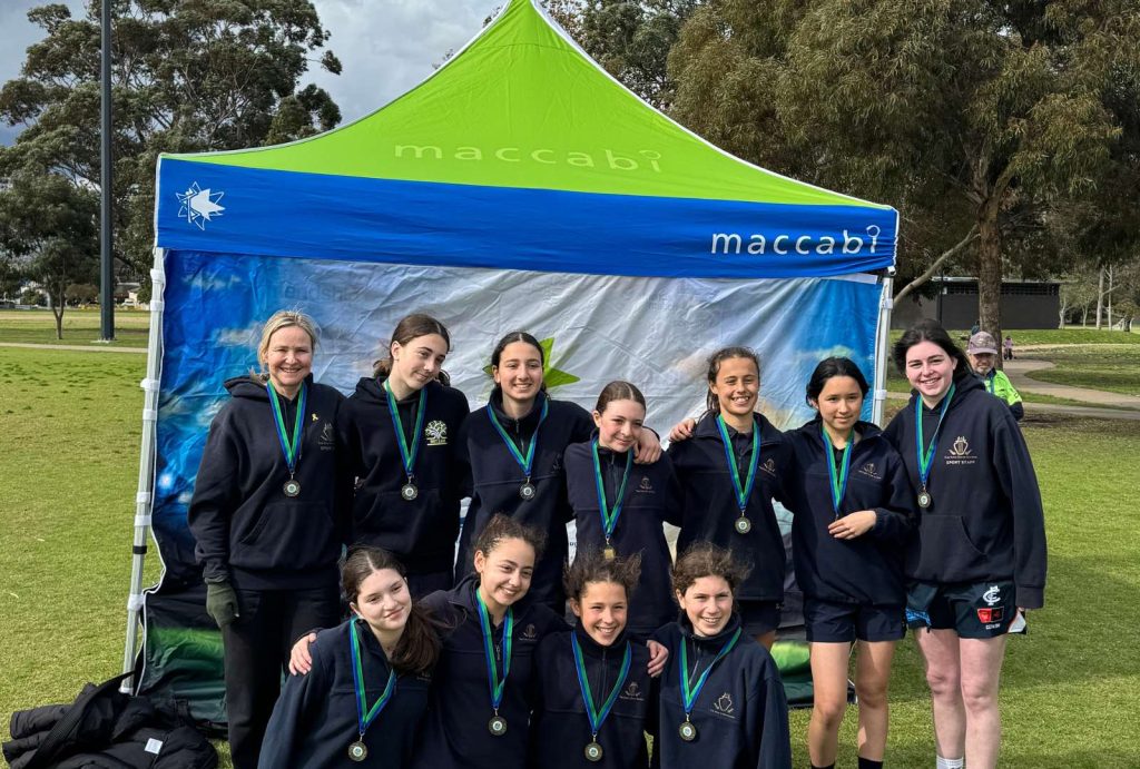The Year 7 Girls' Soccer team are huddled together, smiling at the camera. They are weraing medals. they have just been declared the winners in their division of the Maccabi Soccer Cup.