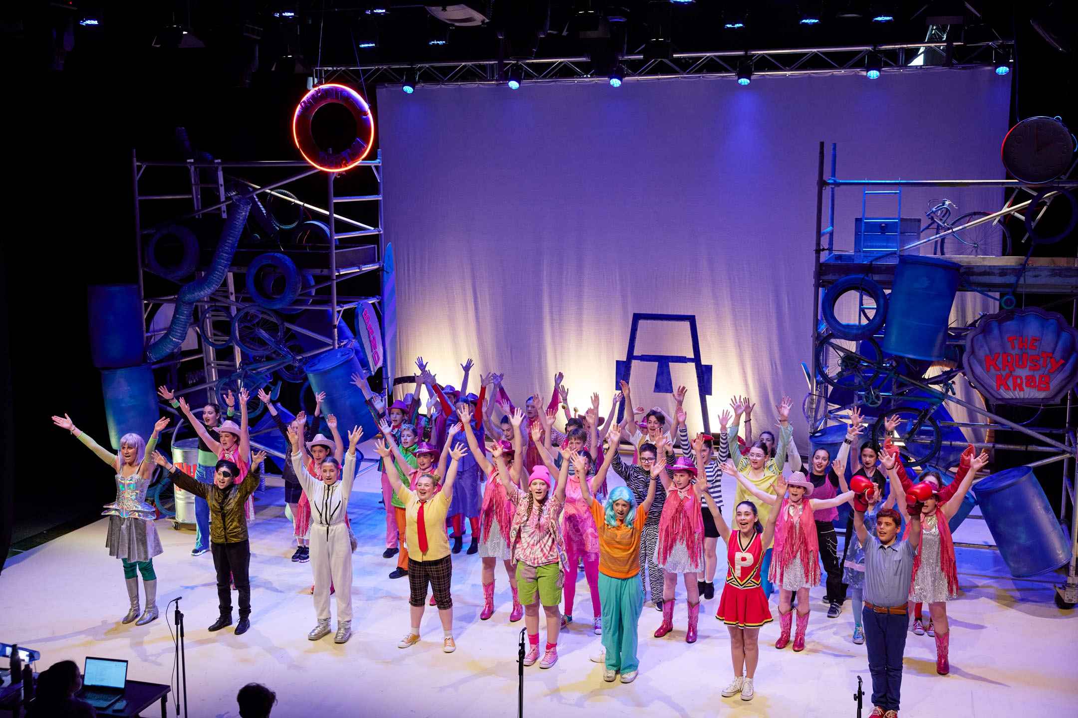 Years 6-9 students are on stage performing in the Middle School Musical, Spongebob. they are in colourful costumes and they are happy.