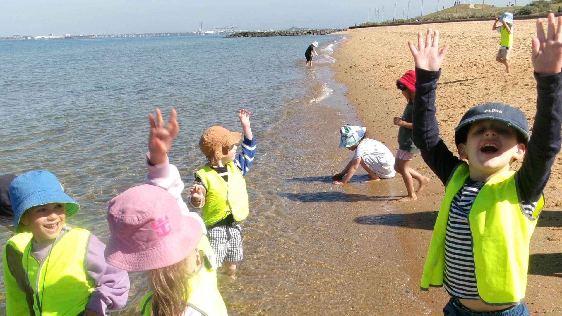 ELC students are playing in the tide. They are wearing neon yellow safety vests and hats. They are touching the water and sand. They are excited to be at the beach.